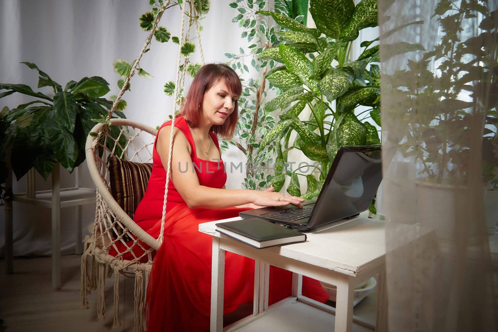 A girl in red dress with laptop is working in chair at a table. A middle-aged business woman. Funny psychologist in the office with flowers