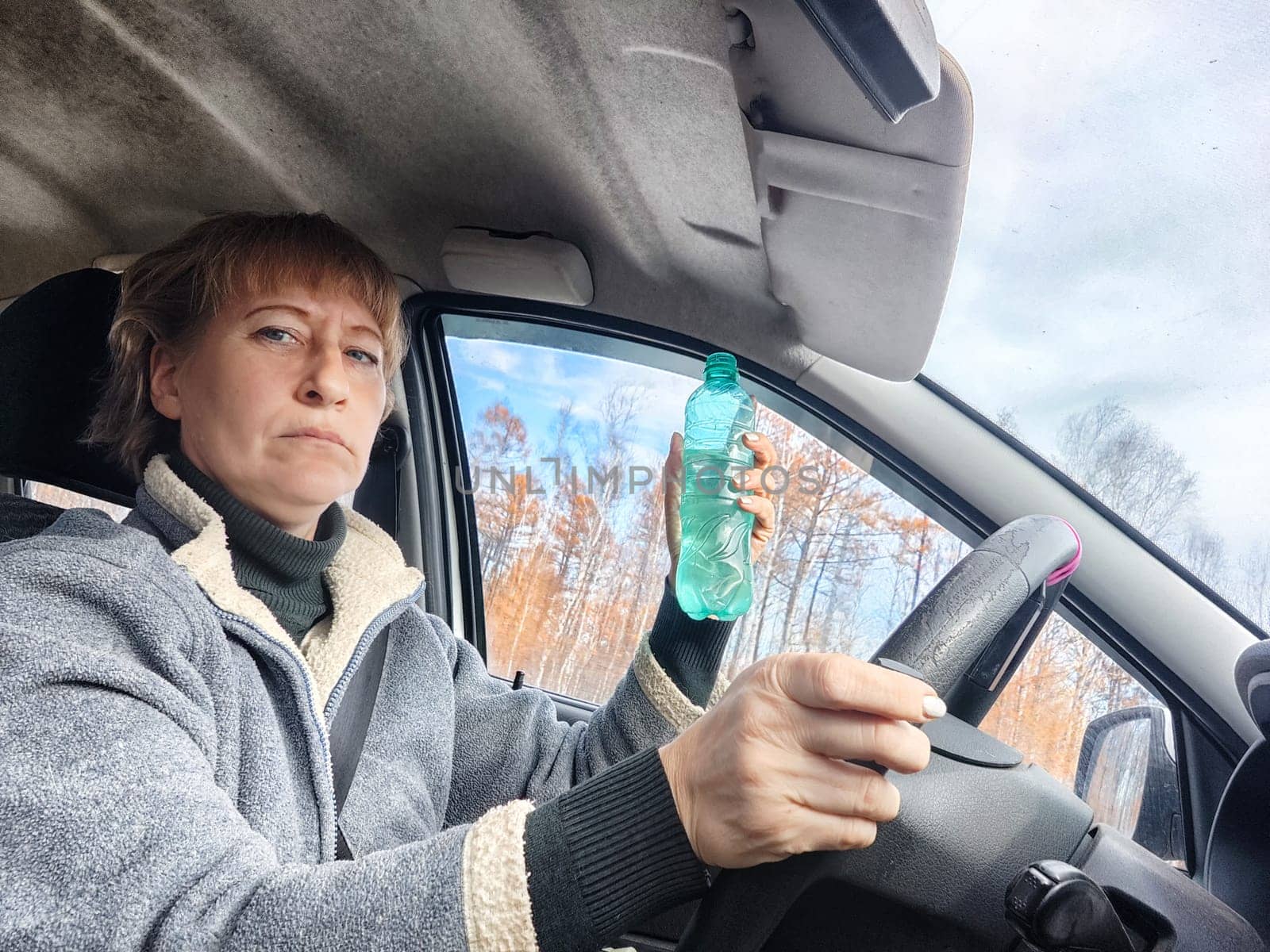 Portrait of female driver in solo journey. Adult mature woman holding steering wheel and bottle of water. Drinking while driving for to stay awake. Lady girl drinks alcohol vodka or gin while driving