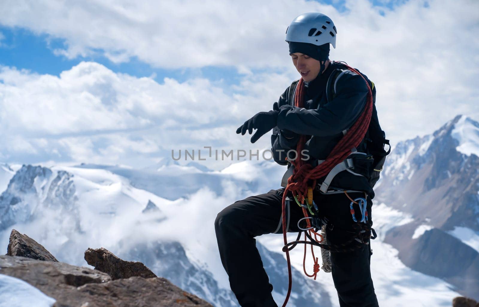 A young man traveler is engaged in mountaineering. Hiker in a helmet, with a rope climbs to the top, against the backdrop of a stunning view with snow-capped mountains.