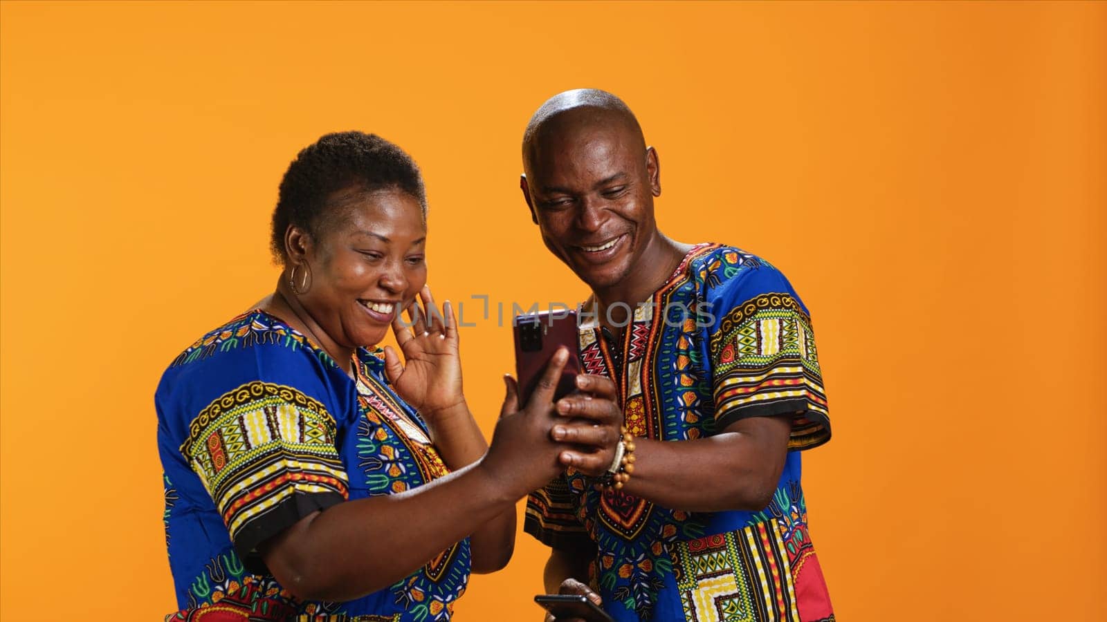 Traditional african american couple browsing websites on smartphone, checking photos on social media app and laughing at memes. Joyful people in colorful attire looking at pictures online.