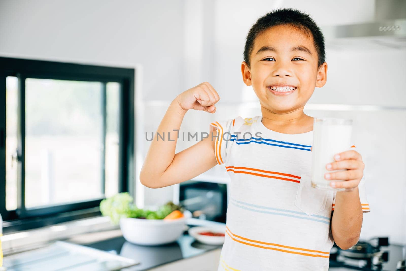 Happy Asian little boy in kitchen holds milk cup, smiling. Portrait of cute son enjoying the drink. Joyful child sips calcium-rich liquid, radiating happiness at home give me. by Sorapop