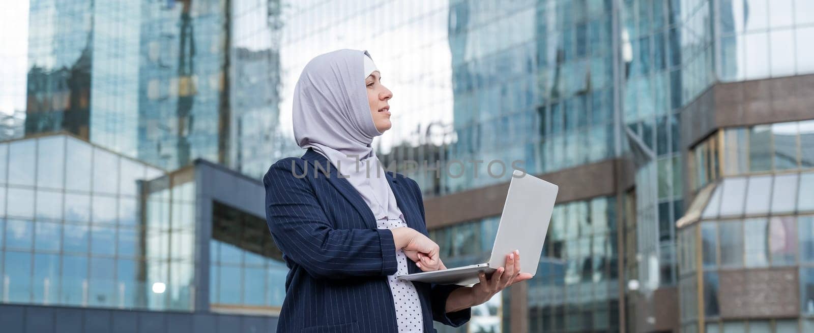 Thoughtful business woman in hijab and suit is holding a laptop outdoors. by mrwed54