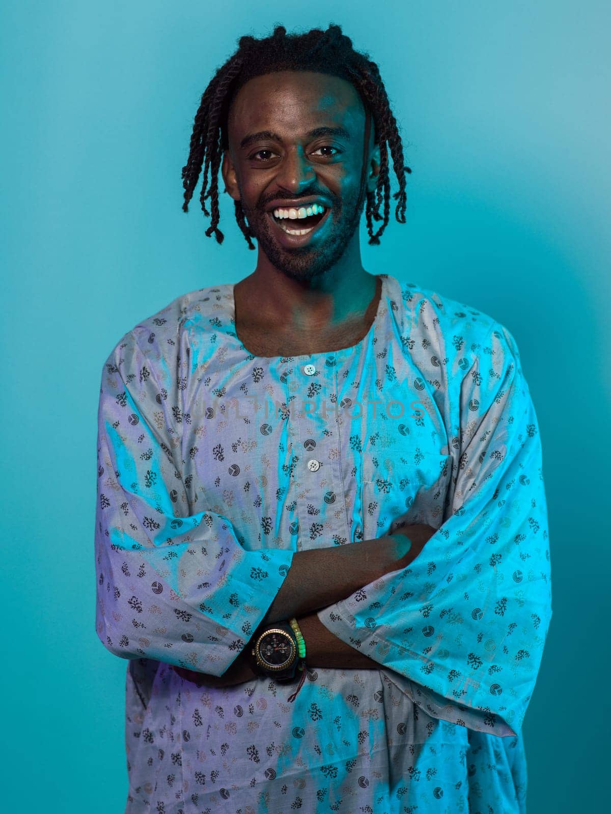 A Sudanese man adorned with modern dreadlocks stands proudly in traditional Sudanese attire, his arms crossed, conveying a blend of cultural heritage and contemporary style against a vibrant blue backdrop. by dotshock