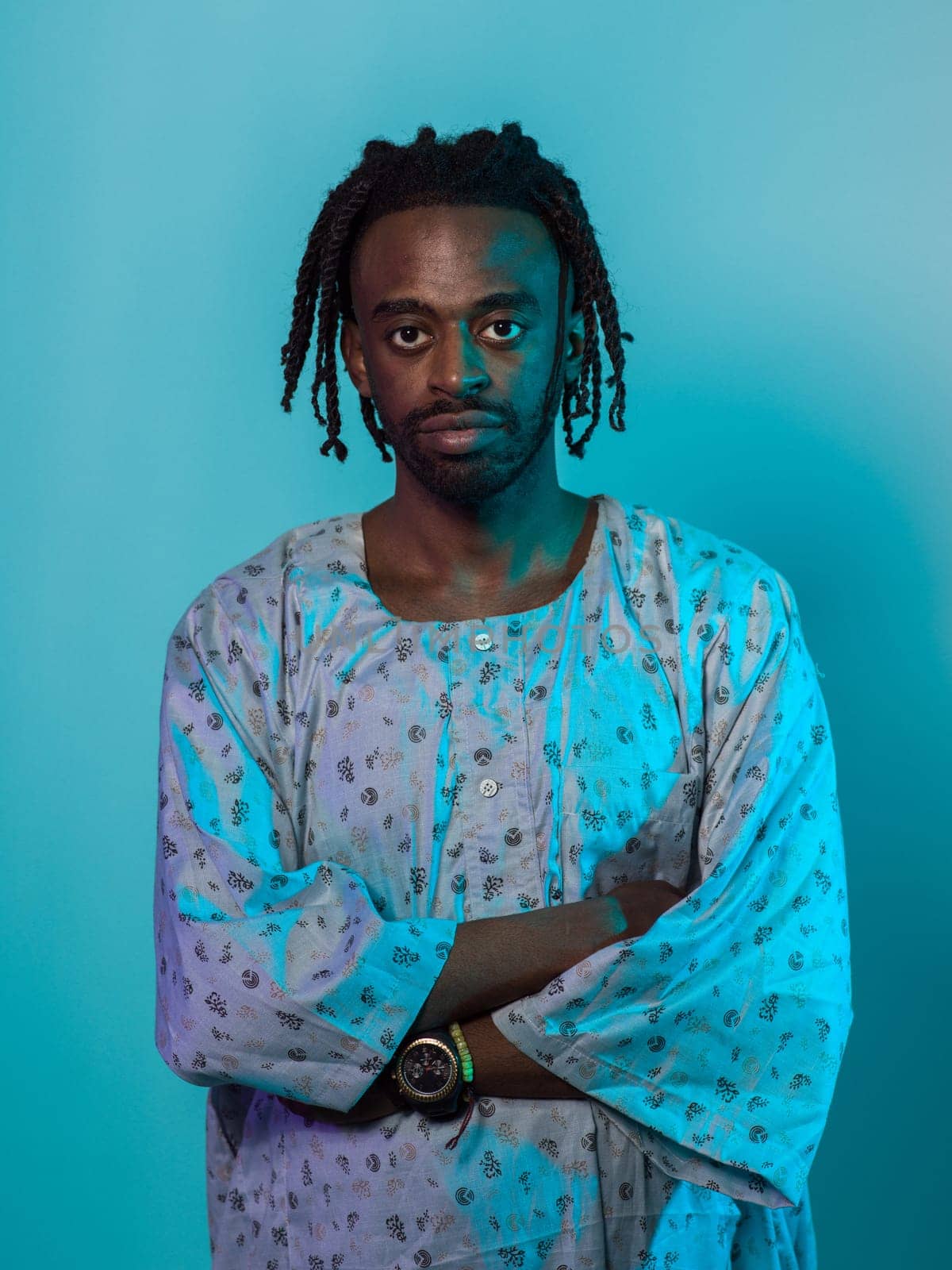 A Sudanese man adorned with modern dreadlocks stands proudly in traditional Sudanese attire, his arms crossed, conveying a blend of cultural heritage and contemporary style against a vibrant blue backdrop. by dotshock