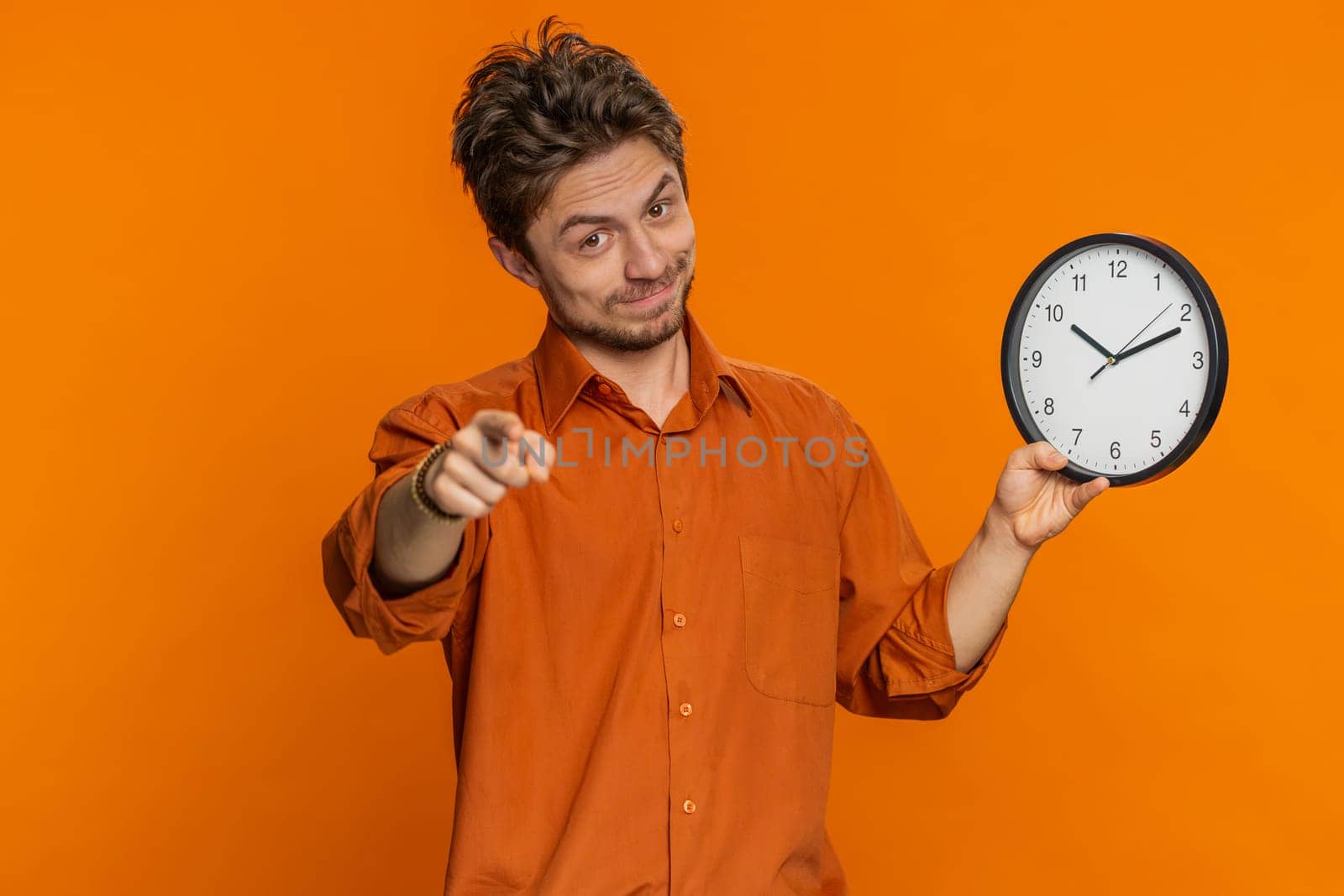 It is your time, hurry up. Caucasian man showing time on wall office clock, ok, thumb up, approve, pointing finger at camera, advertisement, deadline. Young guy isolated on studio orange background