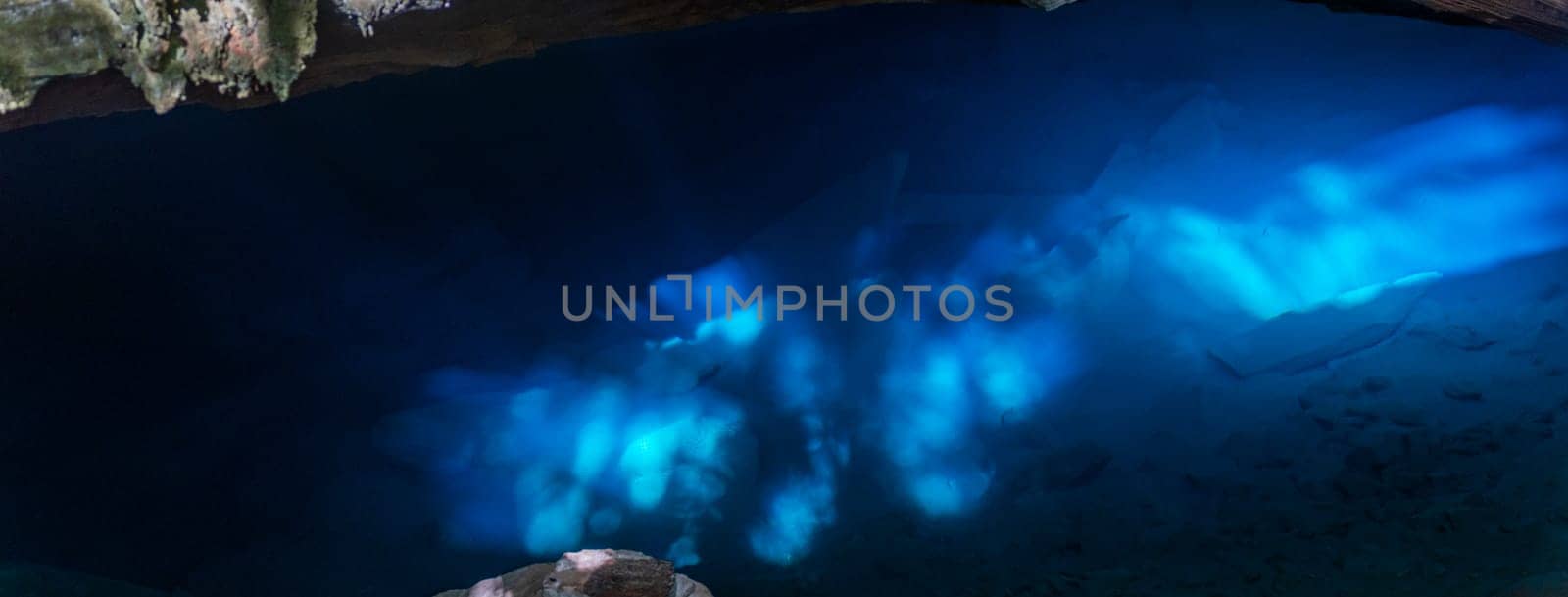 Underwater photo depicts a cave illuminated by surreal blue light, showcasing rocks and silhouetted figures.