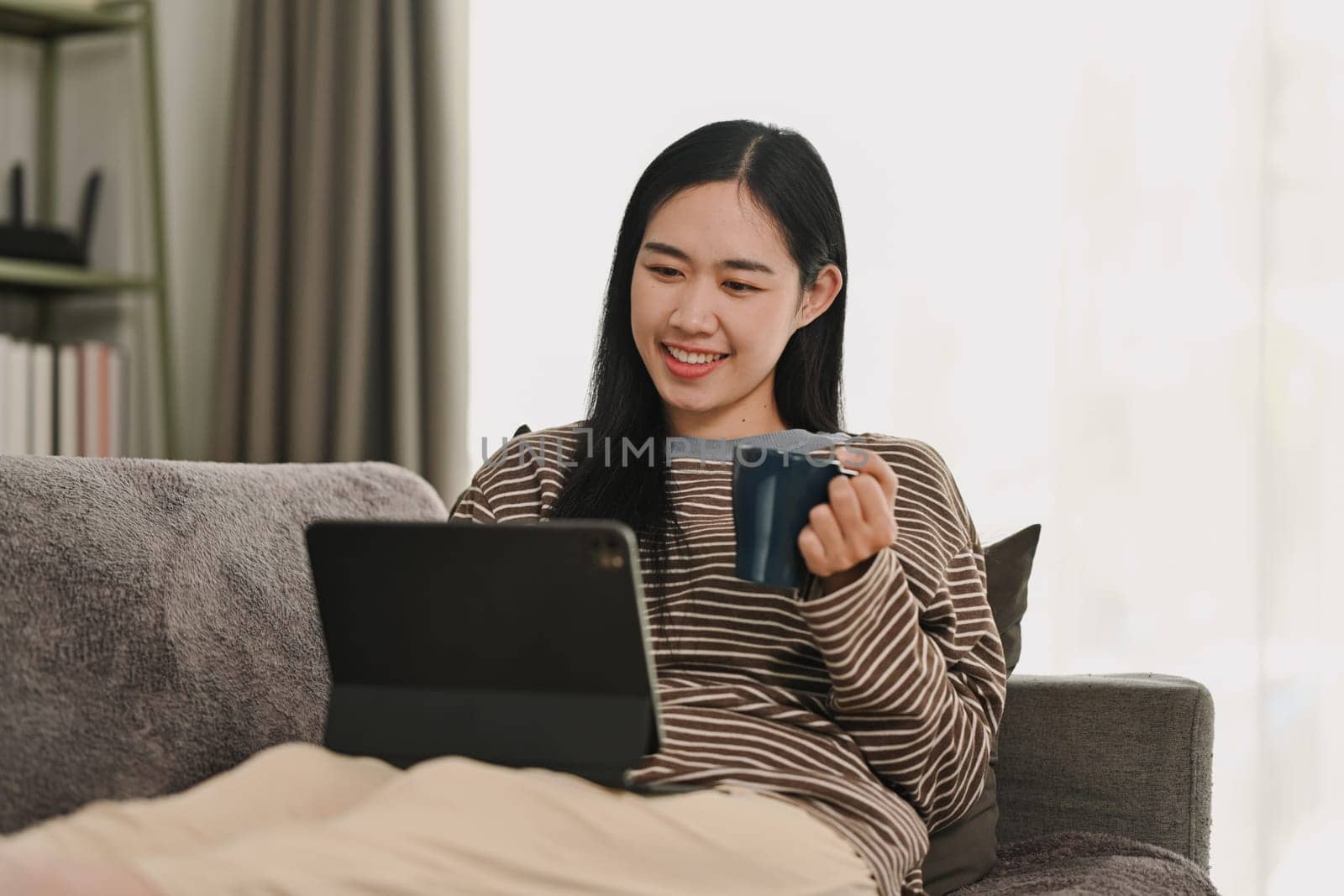 Smiling young woman drinking coffee and working on digital tablet at home.