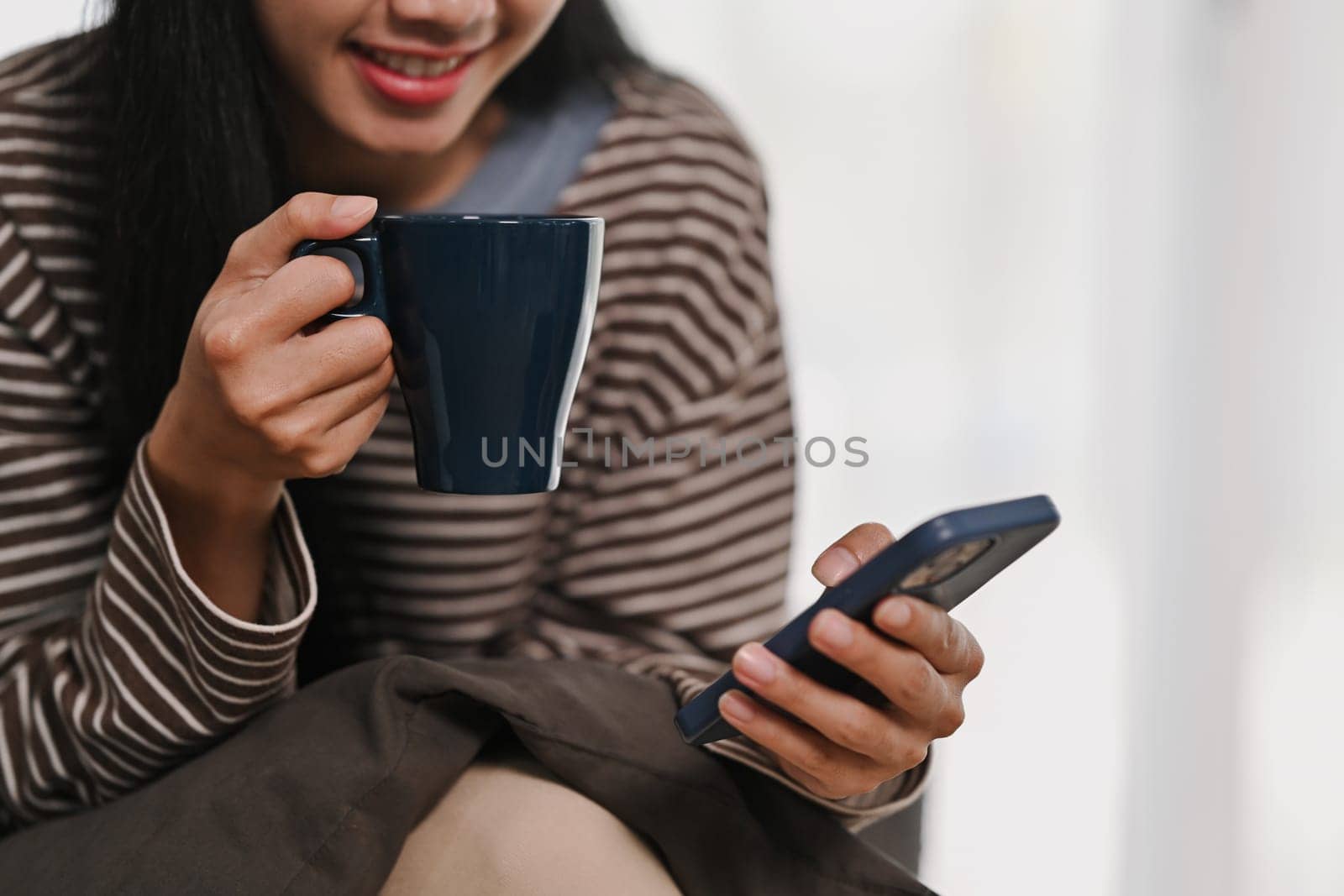 Pleased young woman drinking hot tea and chatting on mobile phone. People and technology concept.