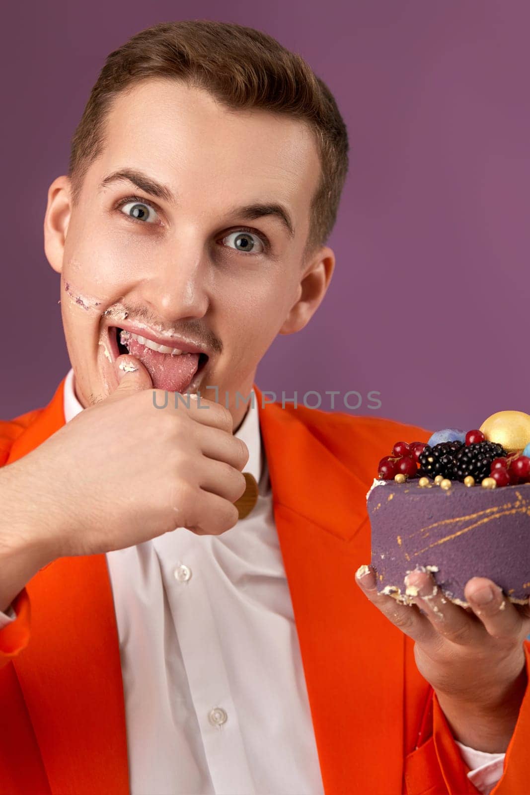 handsome birthday man in orange jacket eating cream cake on purple background