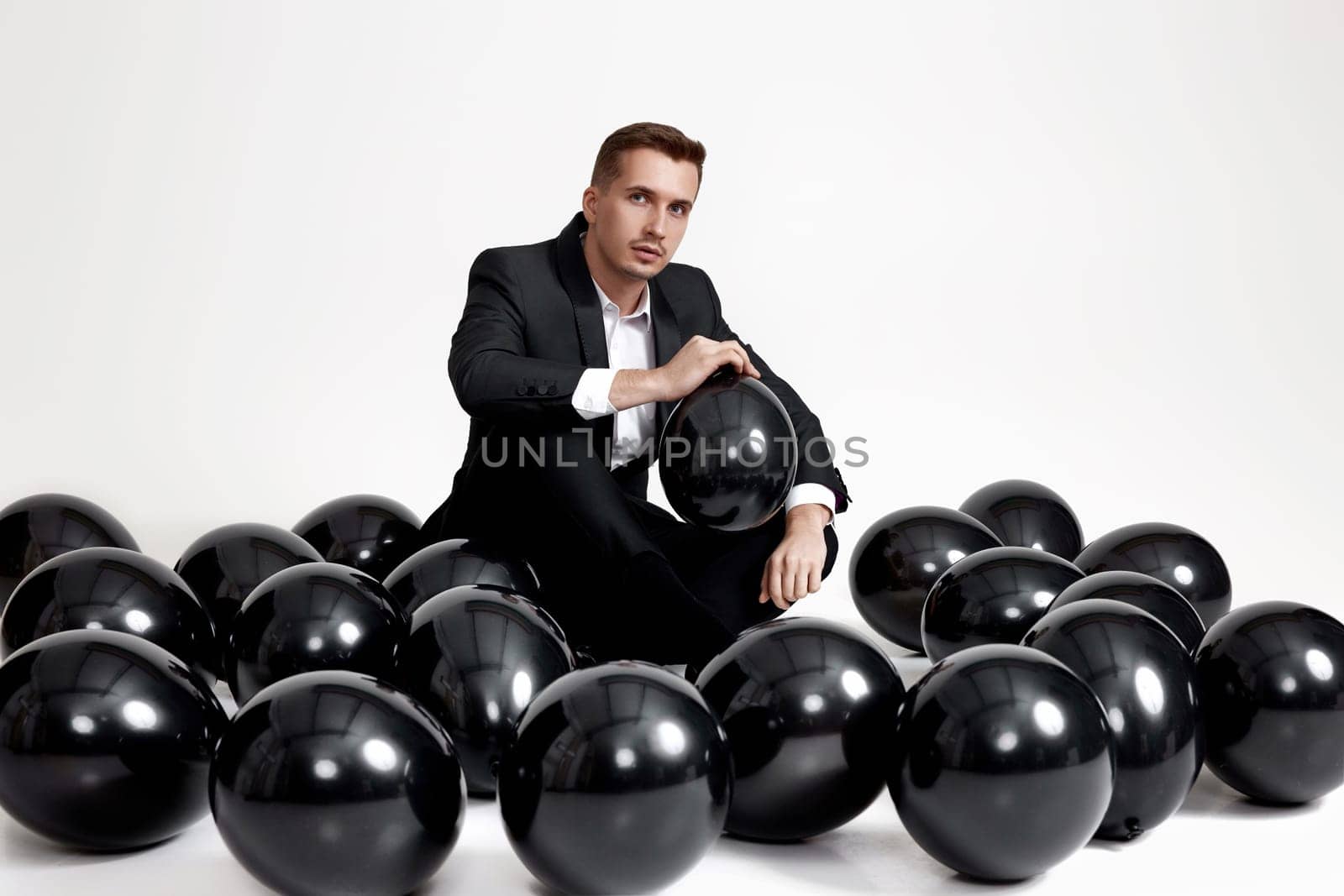 elegant caucasian man in suit tuxedo sitting on studio floor with a lot of black air balloons