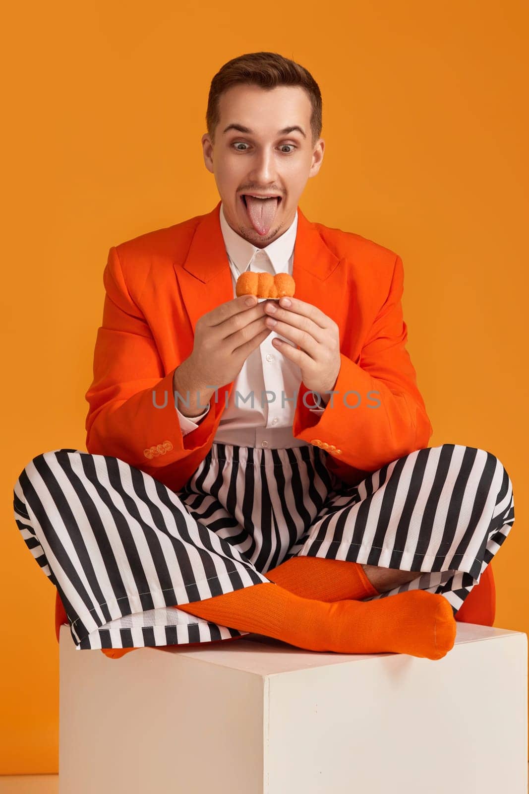 extravagant funny young man in orange jacket and striped pants holding cupcake on orange background.