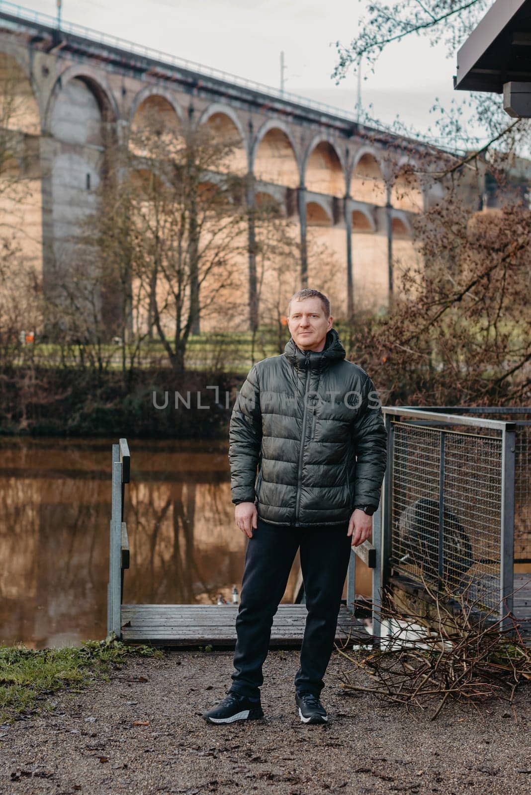 Timeless Elegance: 40-Year-Old Man in Stylish Jacket by Neckar River and Historic Bridge in Bietigheim-Bissingen, Germany. Experience the allure of seasons as a charismatic 40-year-old man stands gracefully by the enchanting Neckar River, adorned in a sophisticated jacket, against the backdrop of historic bridge pillars in the charming city of Bietigheim-Bissingen, Germany. This captivating image seamlessly blends timeless elegance with the scenic beauty of autumn or winter, creating a picturesque scene of urban exploration and mature style in the heart of German heritage