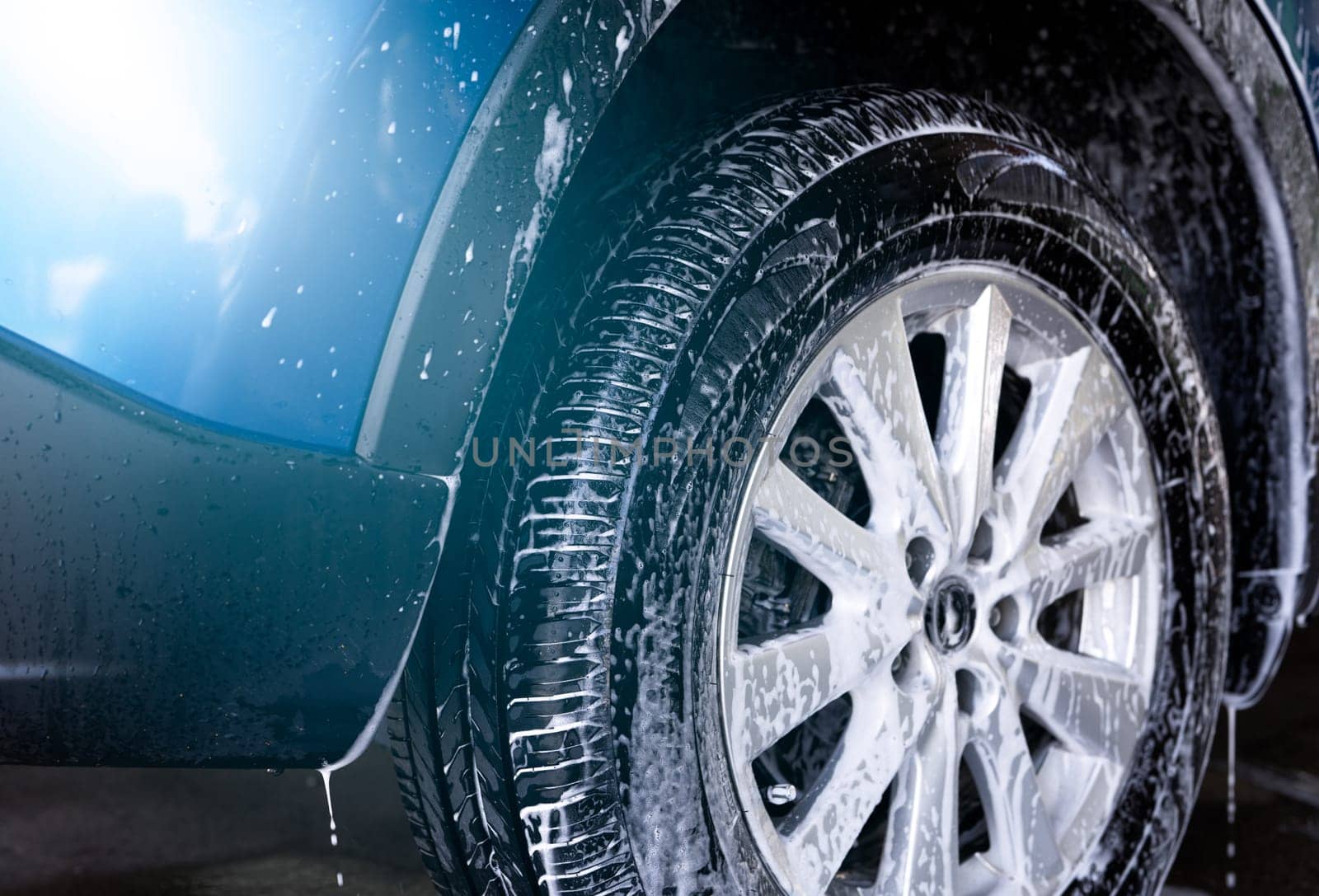 Blue car wash with white soap foam and professional auto care service. Car cleaning service concept. Vehicle cleaning service. Foam wash car detailing. Tire of car wheel is covered with white foam. by Fahroni