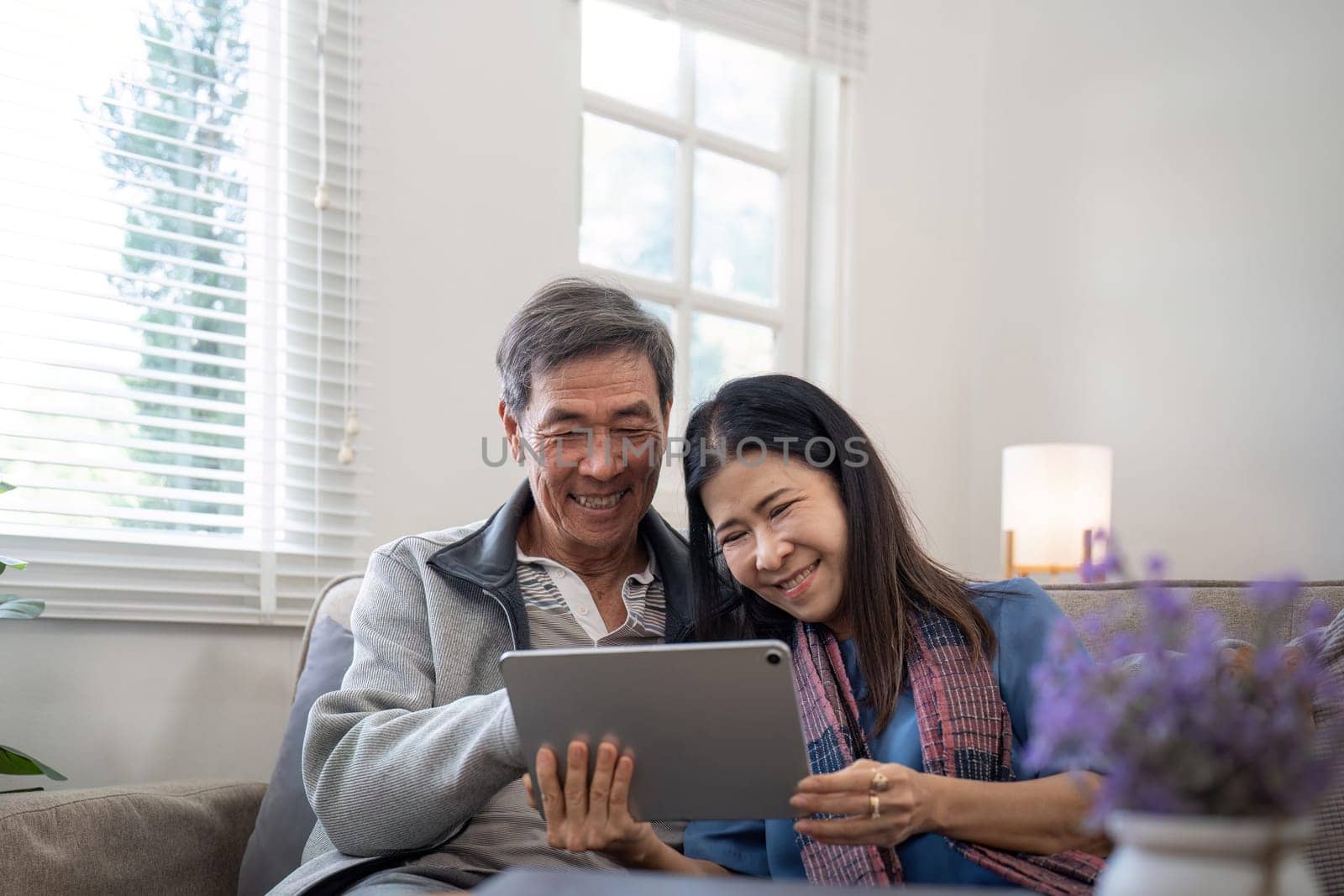 Happy elderly asian couple using tablet sit on sofa doing ecommerce shopping online on website and buying insurance browsing at home.