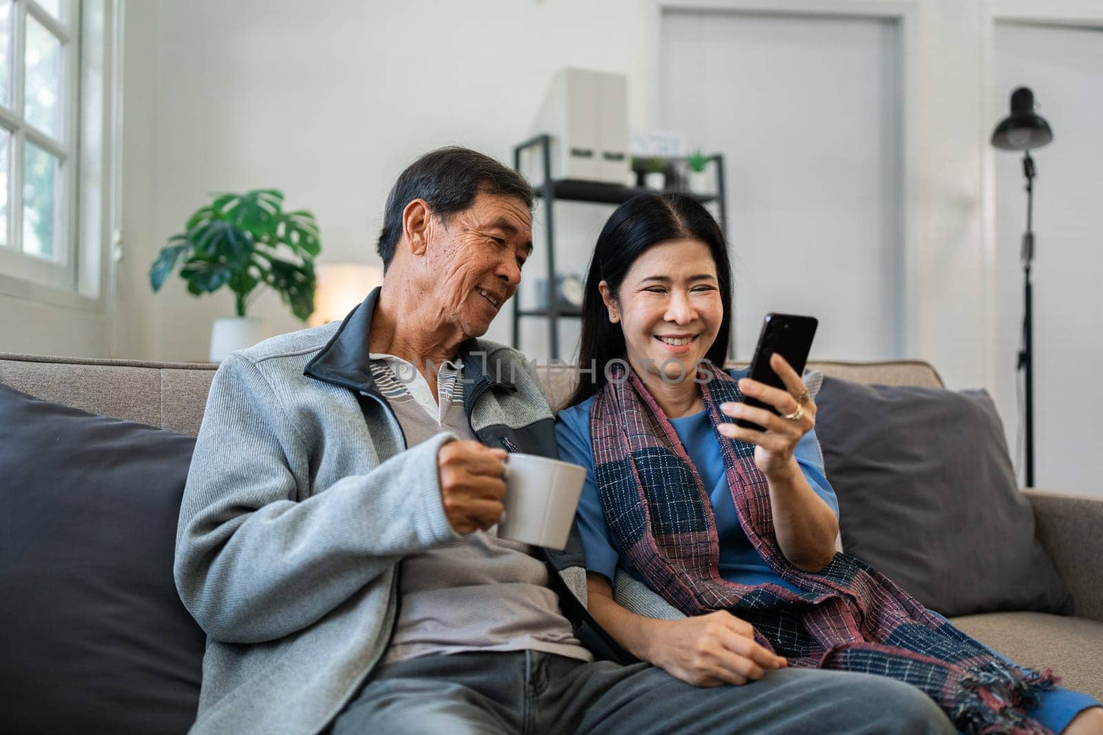 Retired elderly couple sits on couch drink tea and using mobile together and relax in their home. Senior Activity Concept.