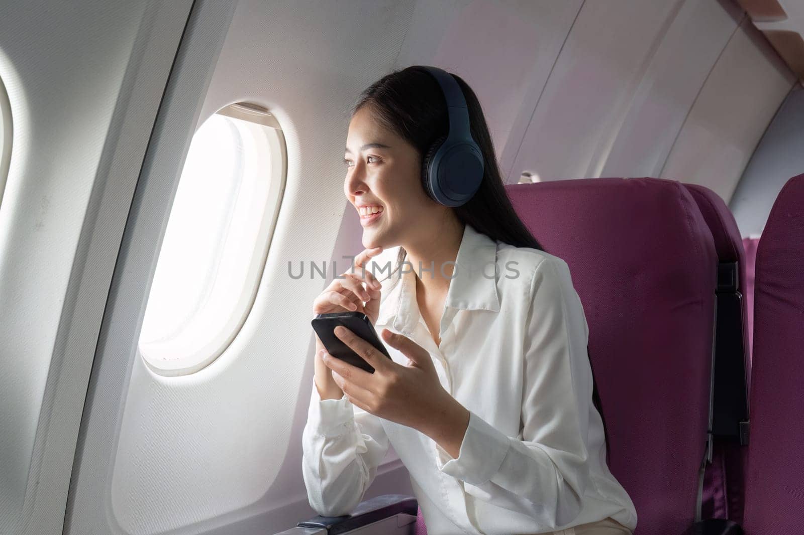 Cheerful Asian woman passenger in headphone watching online movie during intercontinental flight in cabin of aircraft, happy traveler using wifi connection on board by nateemee