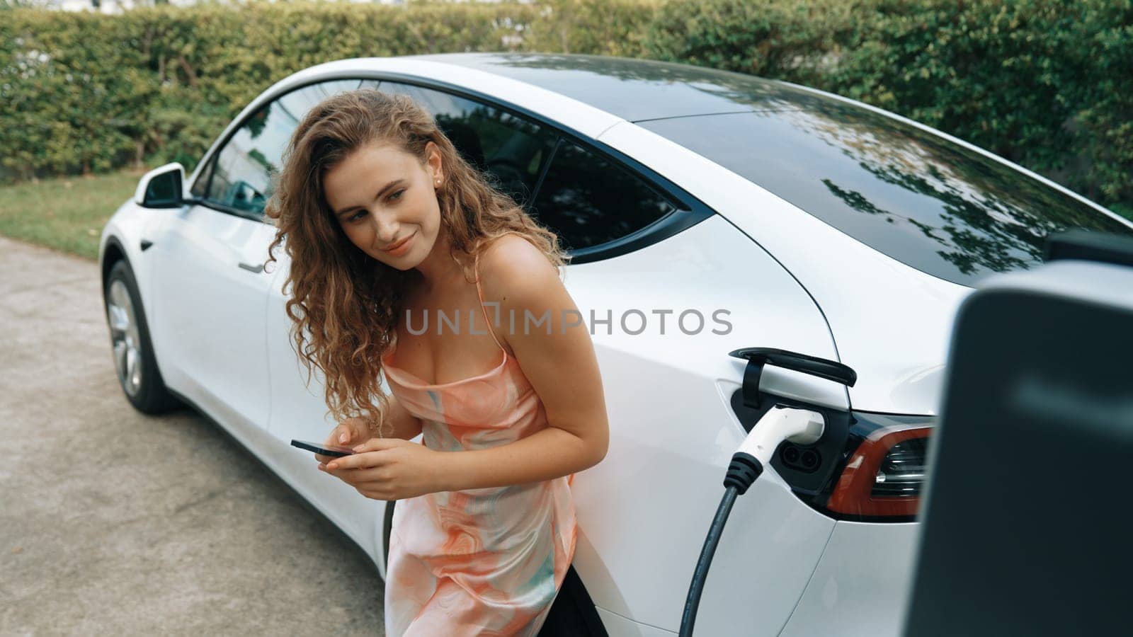 Young woman use smartphone to pay for electricity at public EV car charging station in nature. Modern environmental and sustainable automobile transportation lifestyle with EV vehicle. Synchronos