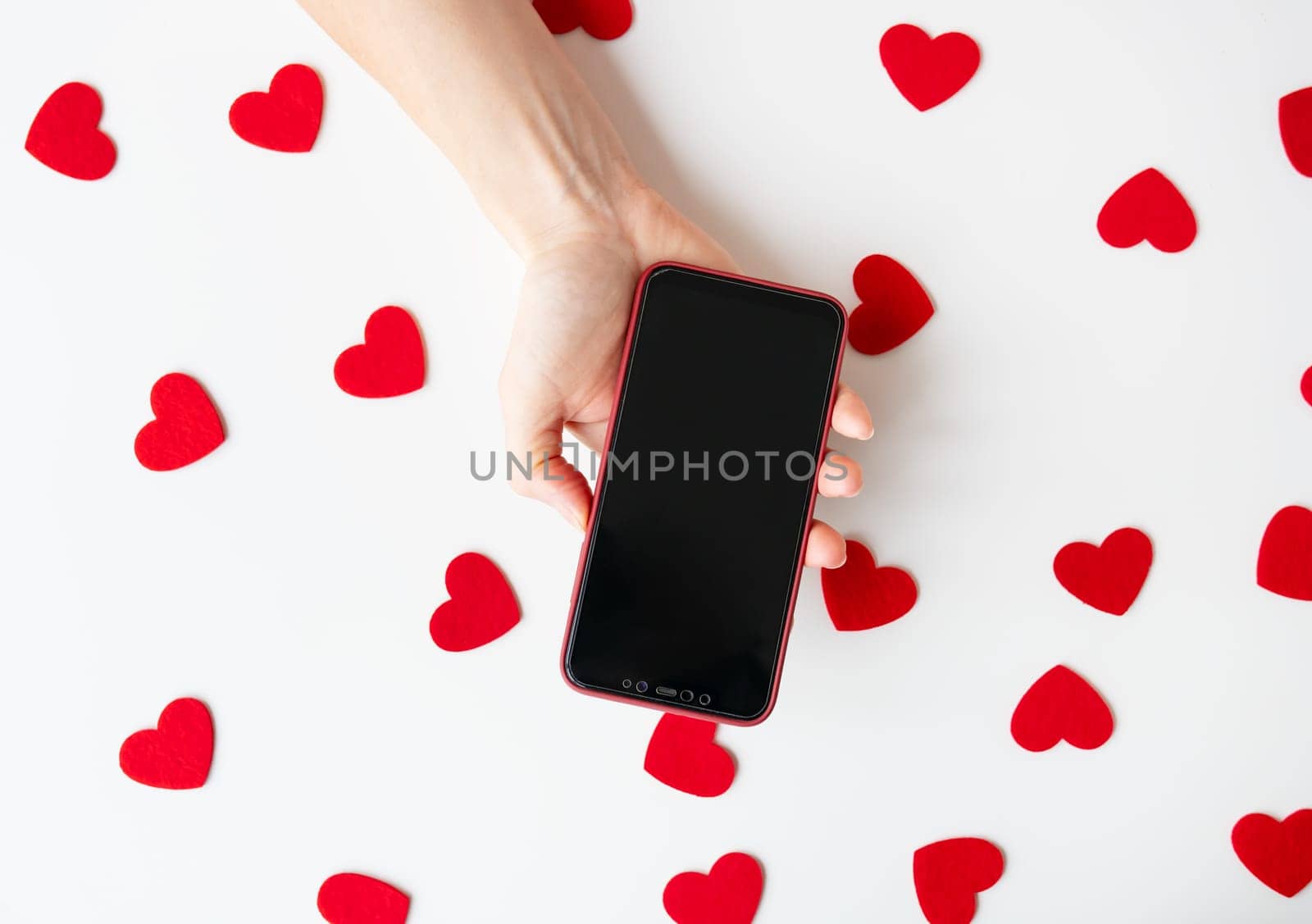 A hand holding a smartphone, surrounded by red heart shapes on a white background. Valentine's Day concept