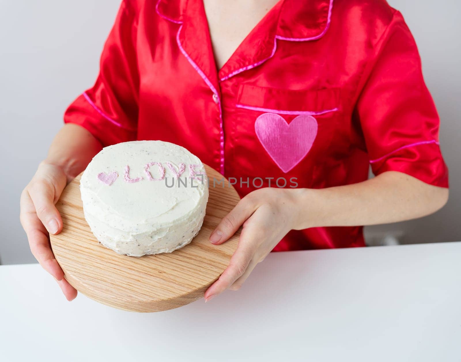 A girl in a red silk shirt with a heart holds a white cake with the inscription LOVE on a wooden board. Preparing for Valentine's Day. by sfinks