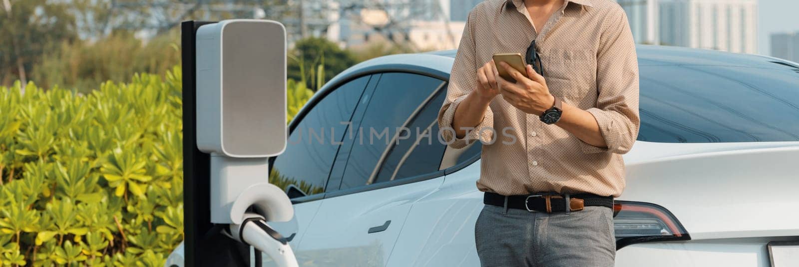 Young man recharge electric car's battery from charging station in outdoor green city park. Rechargeable EV car for sustainable environmental friendly urban travel. Panorama Expedient