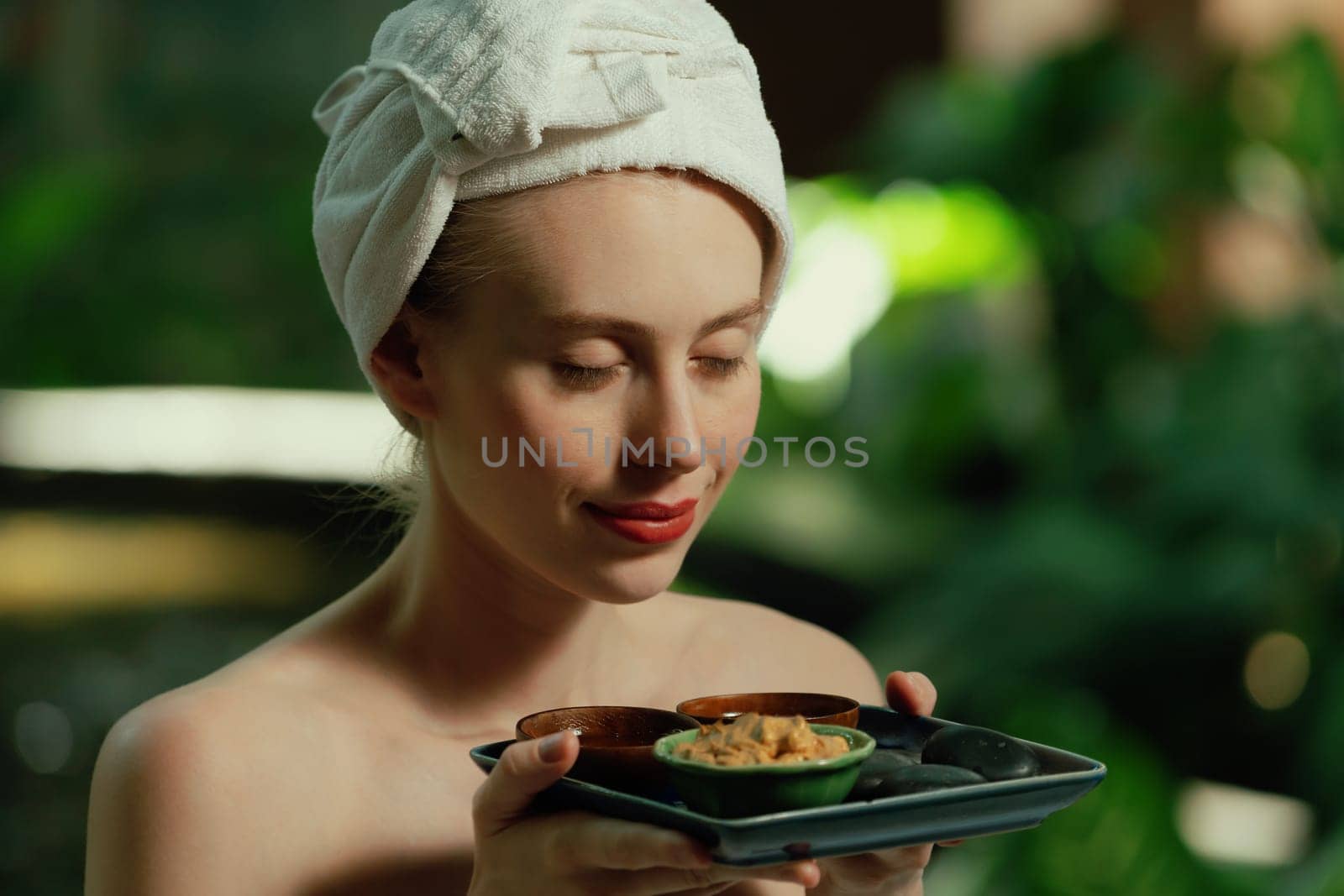 Portrait of beautiful caucasian woman in white towel holds the tray which contained the bowl of herbal scrub and spa rock surrounded by calm and relaxing nature. Side view. Tranquility.