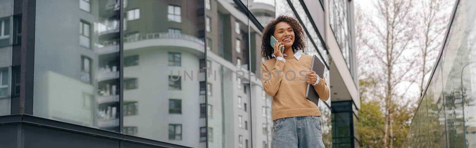 Smiling woman manager is talking phone while walking with laptop on modern building background