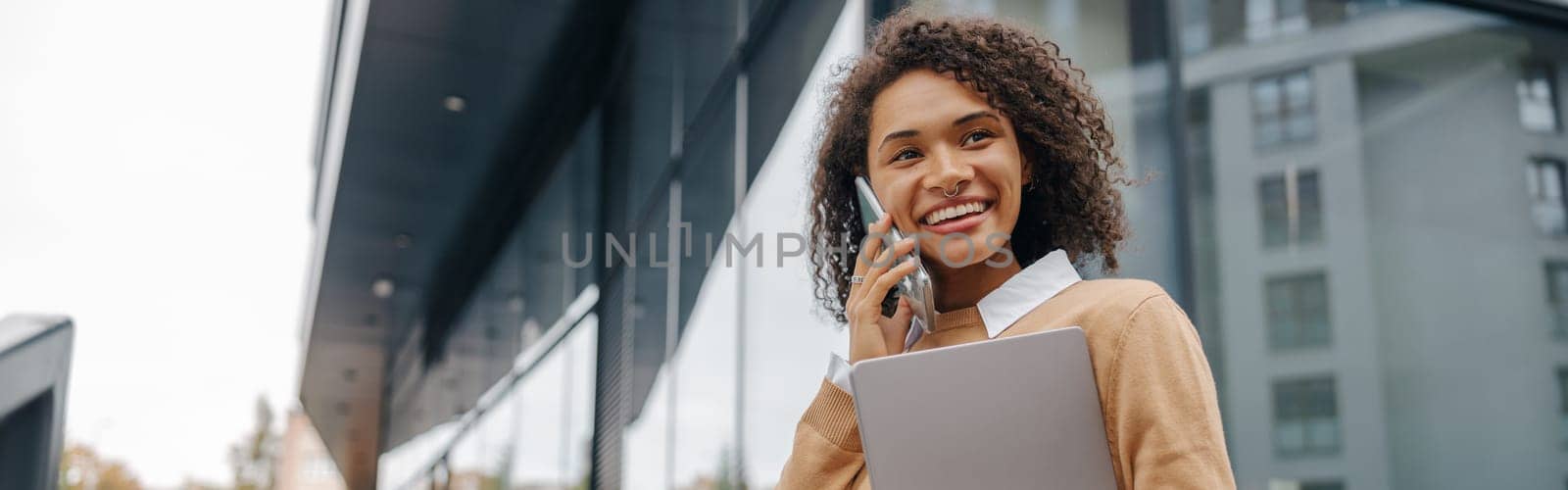 Pretty female entrepreneur is talking phone while standing with laptop on modern building background