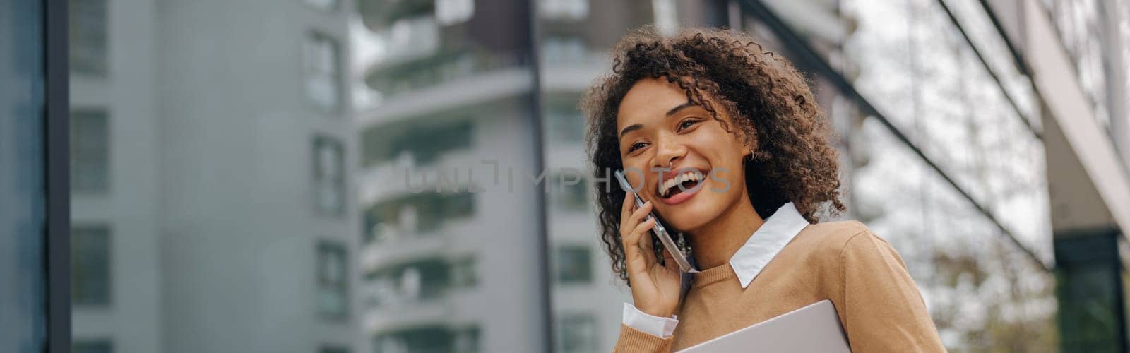 Smiling woman manager is talking phone while standing with laptop on modern building background by Yaroslav_astakhov