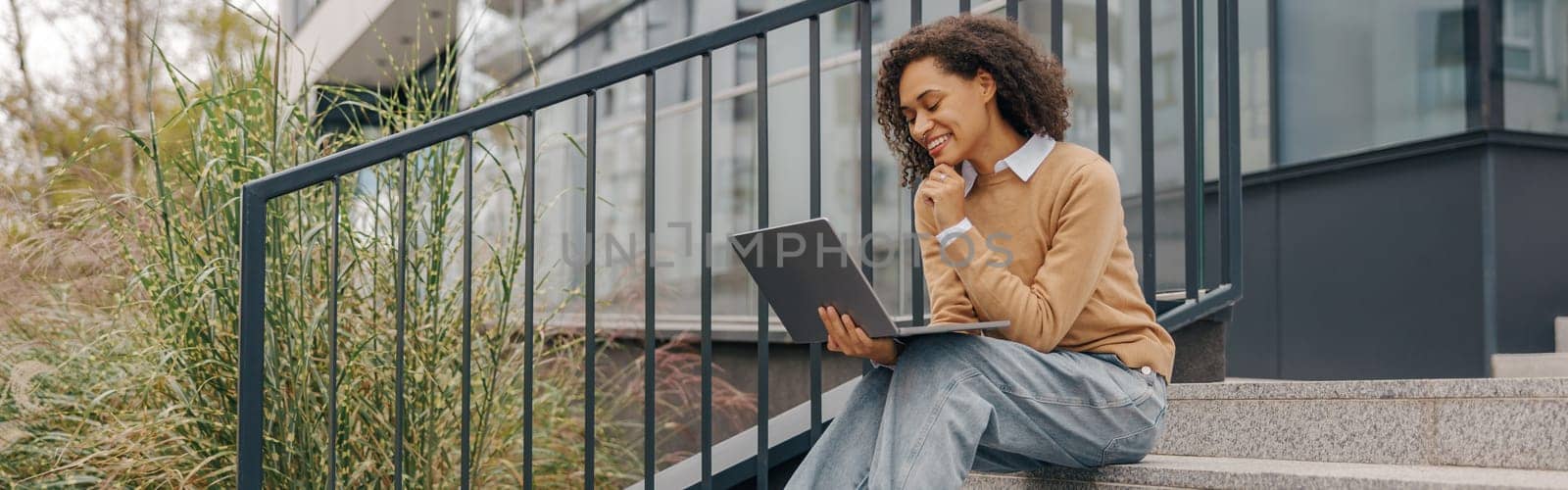 Smiling woman manager working laptop sitting on stairs on modern building background