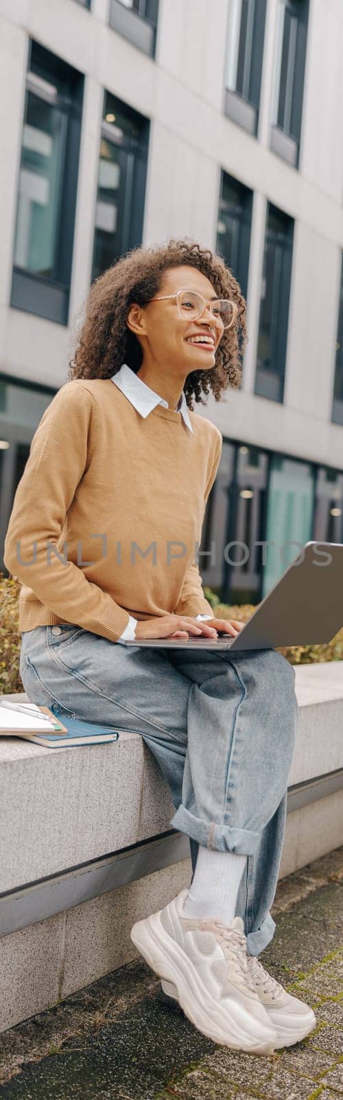 Pretty female entrepreneur working on laptop sitting outside on modern building background