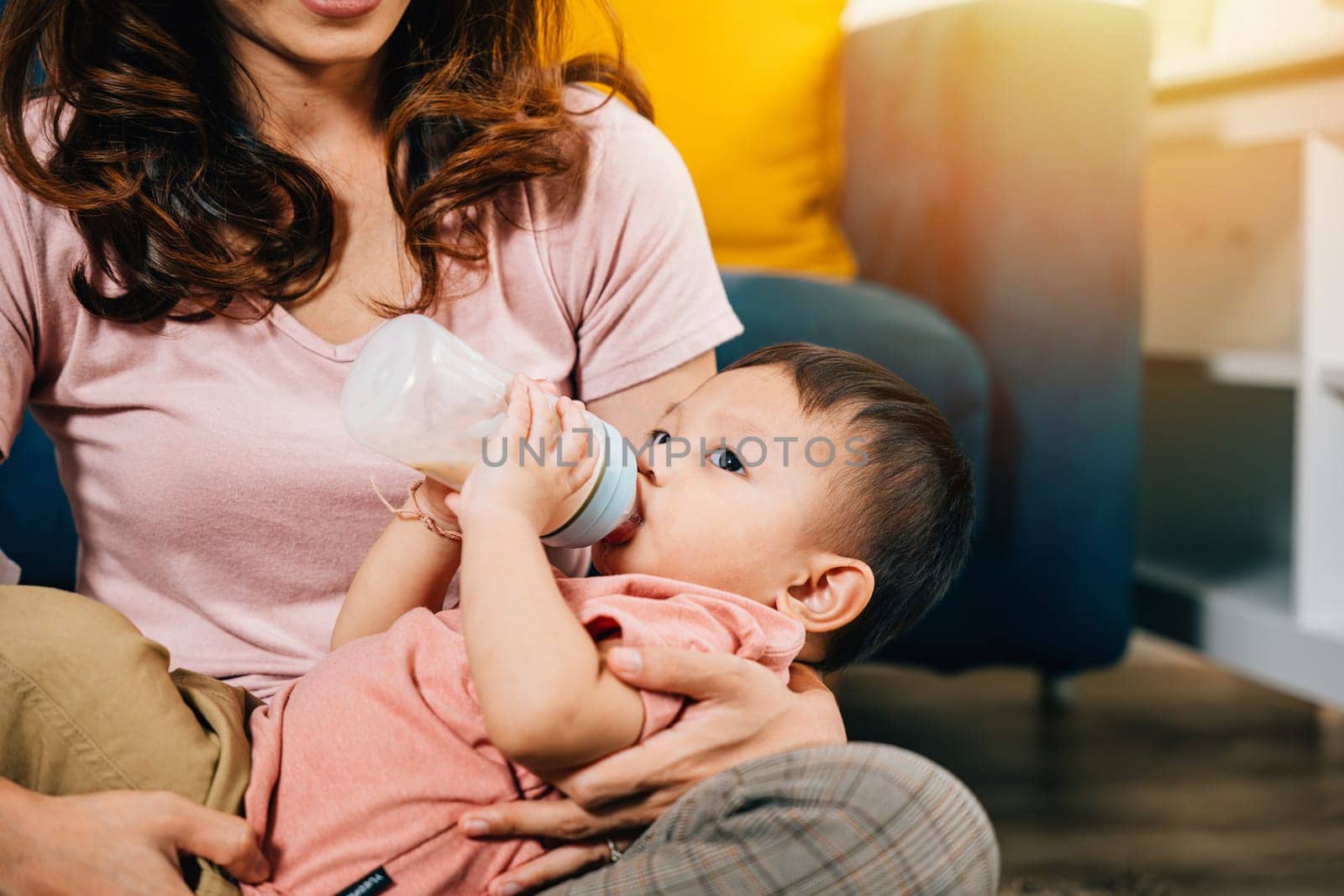 A mother in the cozy living room at home holds her cute Asian baby who is feeding from a baby bottle by Sorapop