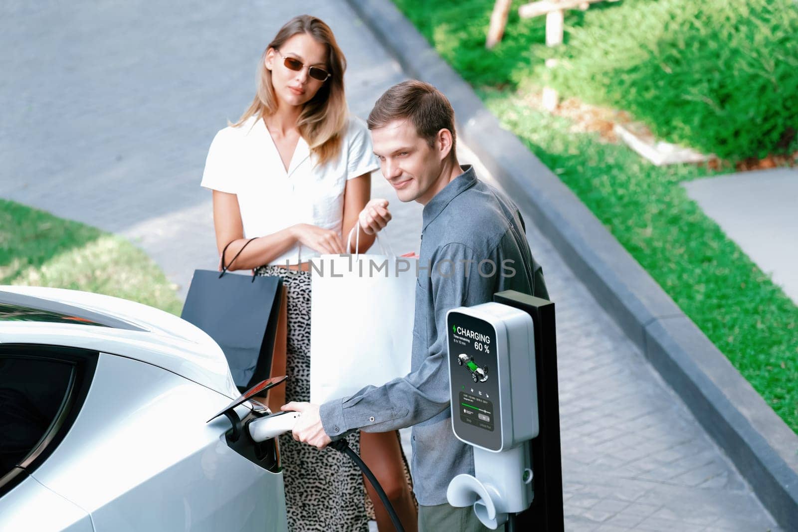 Young couple travel with EV electric car charging in green sustainable city outdoor garden in summer shows urban sustainability lifestyle by green clean rechargeable energy of electric vehicle innards