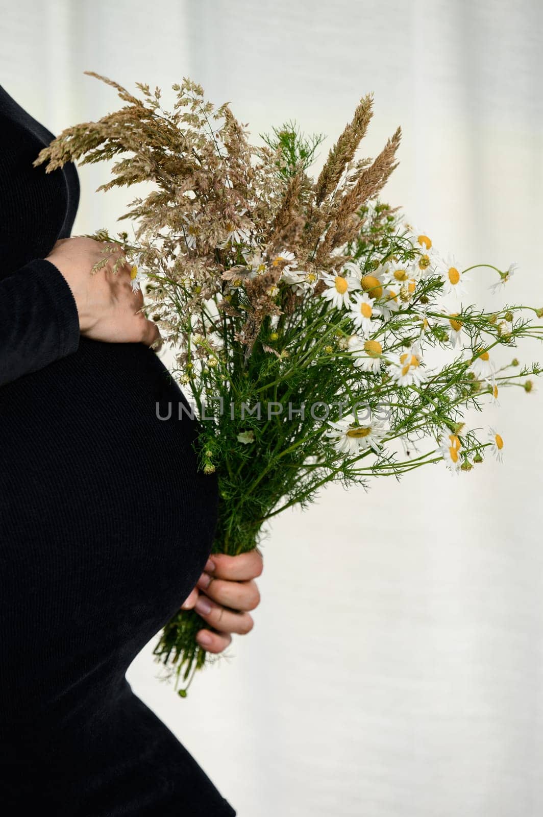 A pregnant woman is hugging her belly against a white background and wearing a black bodycon dress and holding a bouquet of wildflowers in one hand.