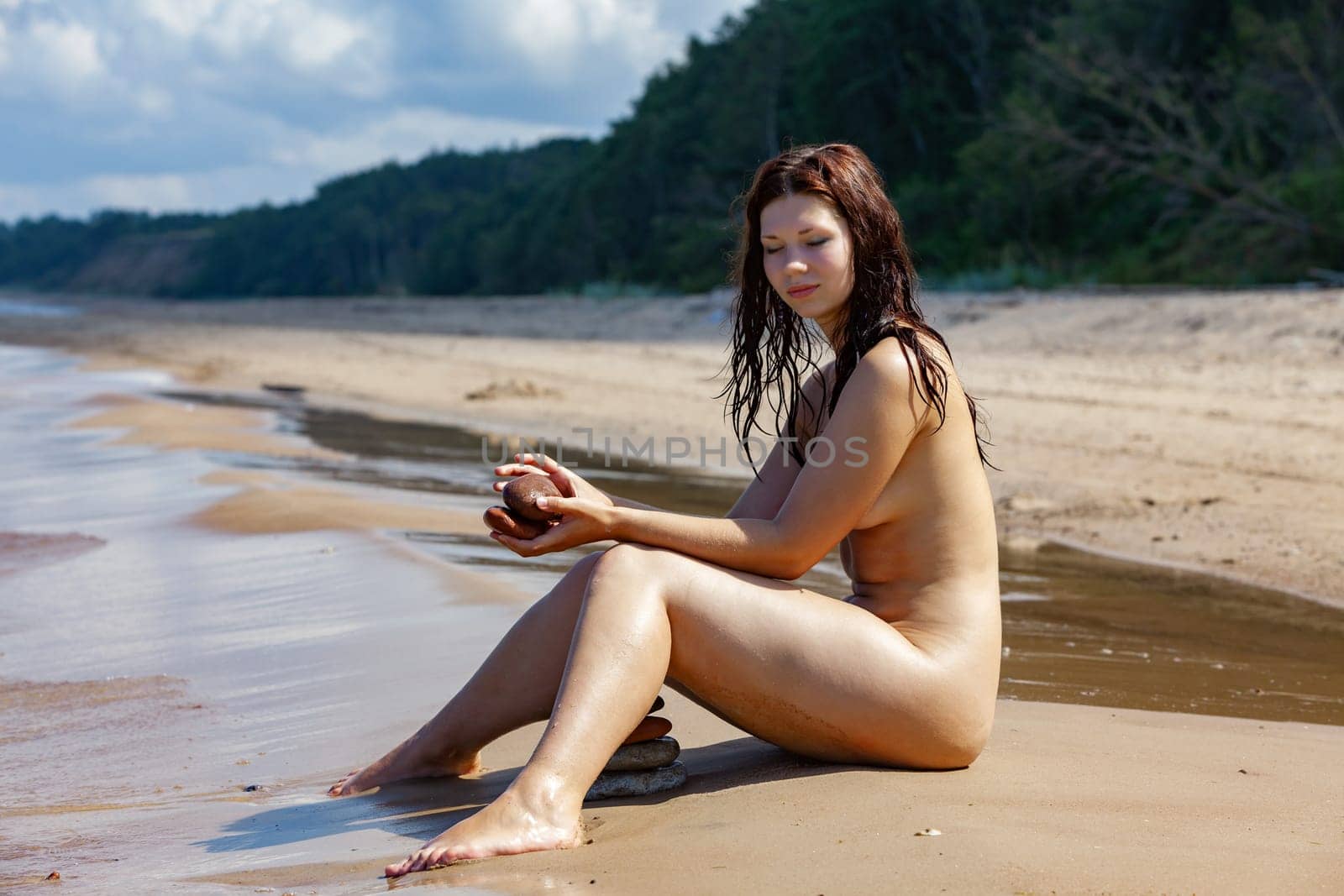 A young completely naked woman sunbathing on the beach, enjoying nature and solitude, building a pyramid out of small stones.
