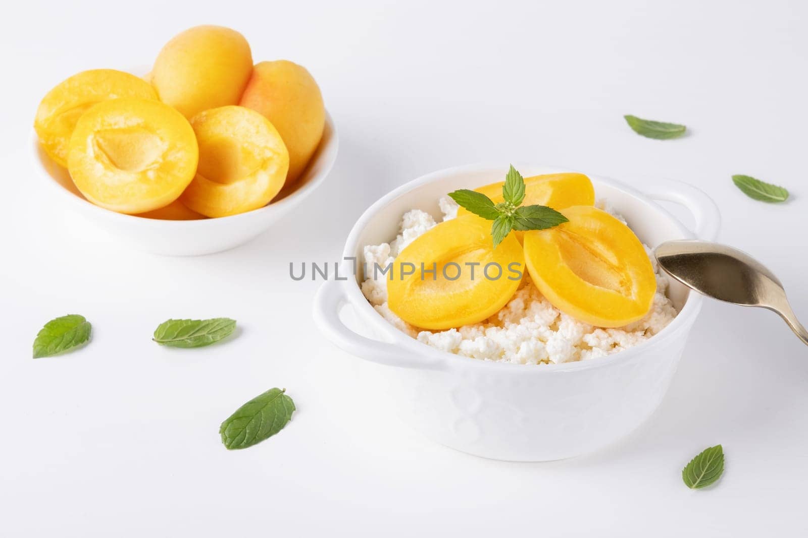 Fresh cottage cheese in white bowl with mint leaves and apricot pieces on white background.