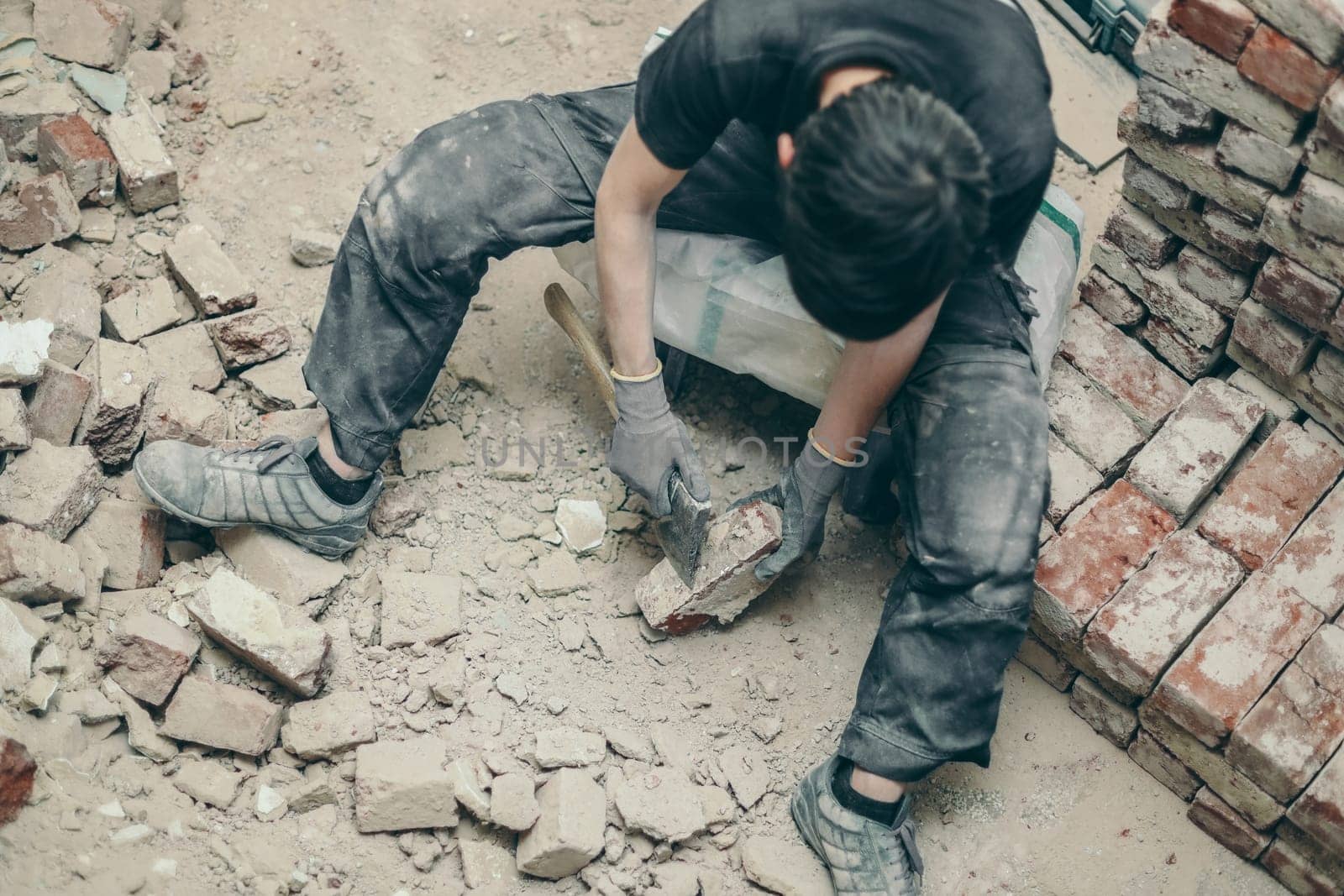 A young caucasian brunette man in black work clothes sits on a small chair and cleans the bricks from dirt and old cement with an ax, top view close-up.Construction work concept.
