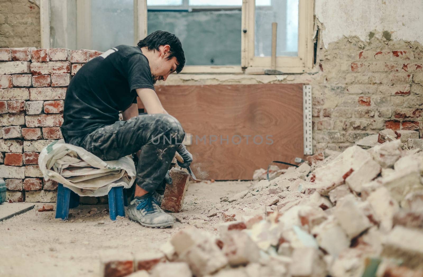 A young Caucasian brunette man with a beard in black work clothes sits sideways on a small chair on the left and cleans the bricks from dirt, debris and old cement with an ax, close-up side view.Construction work concept.