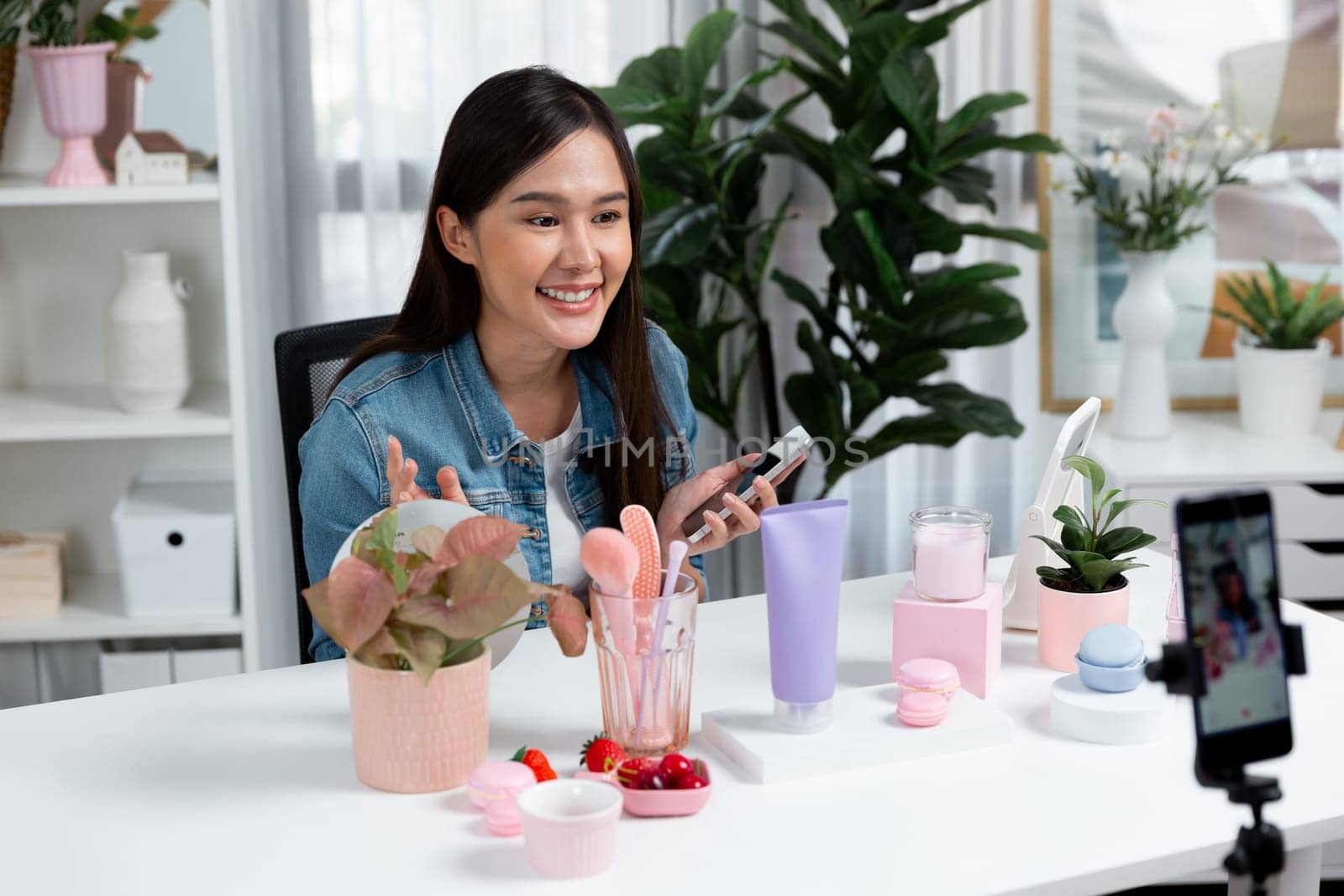 Young beautiful Asian reviewing cosmetic makeup and skin care collection, talking customer on social media online, presenting product surrounded beauty merchandise recording blurred phone . Stratagem.