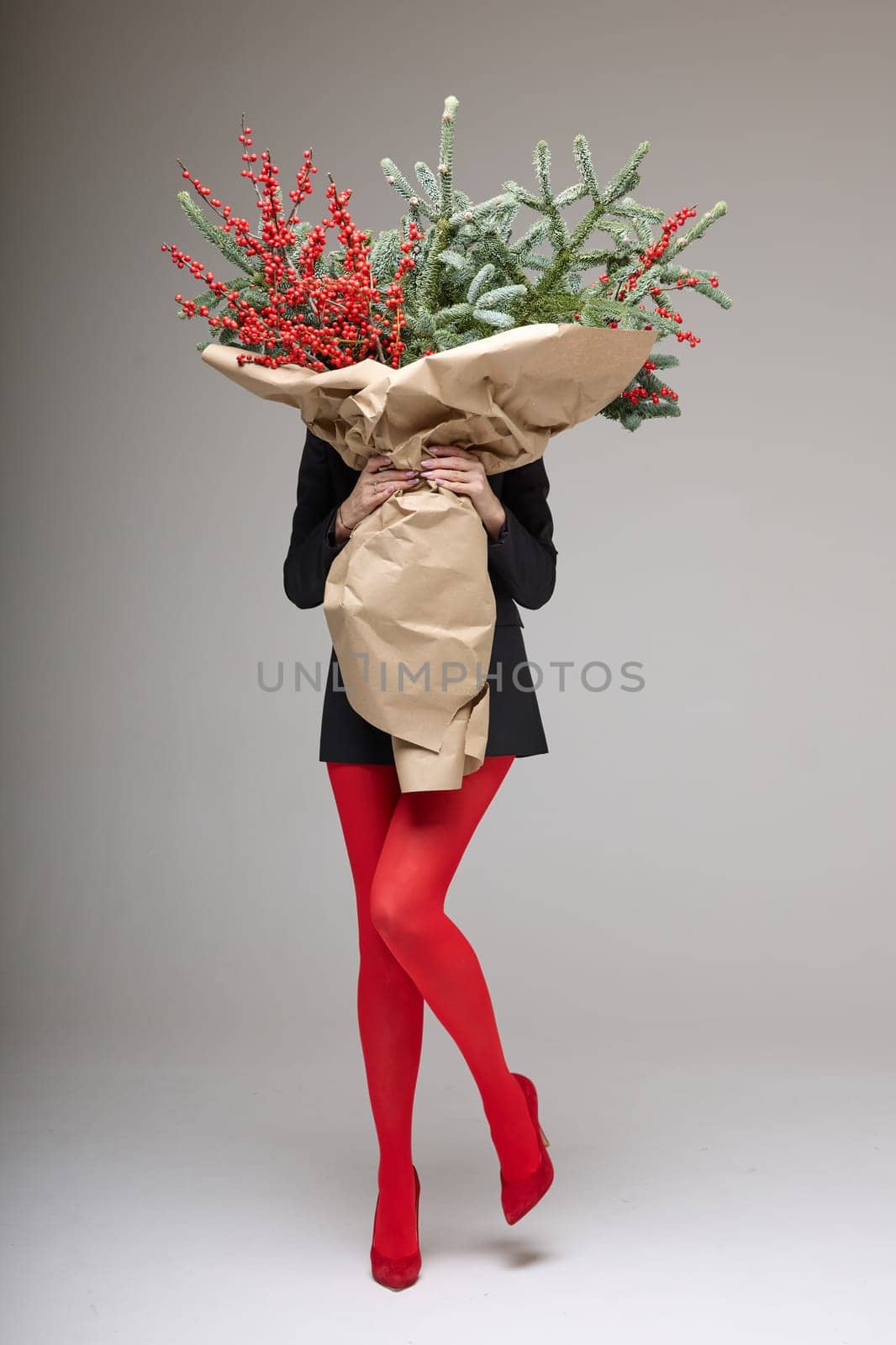 A long girl in red tights and high-heeled shoes holds a huge bouquet of fir branches and red berries wrapped in cardboard paper, she closes with a bouquet, photo studio with white background by vladimirdrozdin