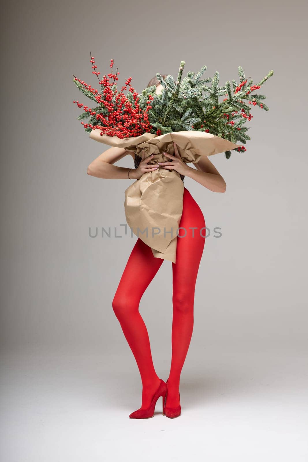 A long girl in red tights and high-heeled shoes holds a huge bouquet of fir branches and red berries wrapped in cardboard paper, she closes with a bouquet, photo studio with white background. High quality 4k footage