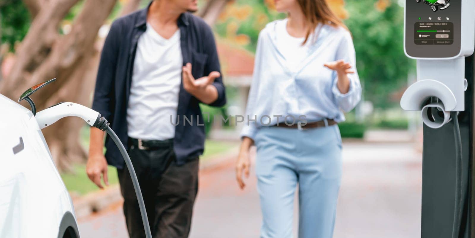 Focused EV car recharging battery on blurred background of lovey couple during autumnal road trip vacation by electric vehicle recharging battery. Eco friendly travel during autumn. Panorama Exalt