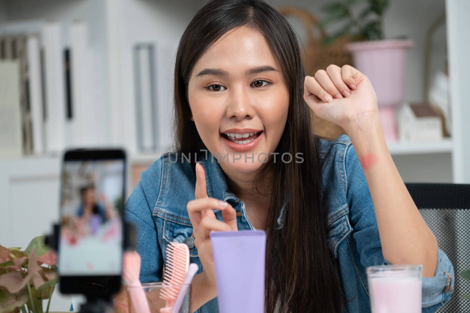 Young beautiful Asian showing cosmetic makeup lipstick matte pastel color testing in bestseller collection on arm test promotion on social media online recording smartphone at modern room. Stratagem.