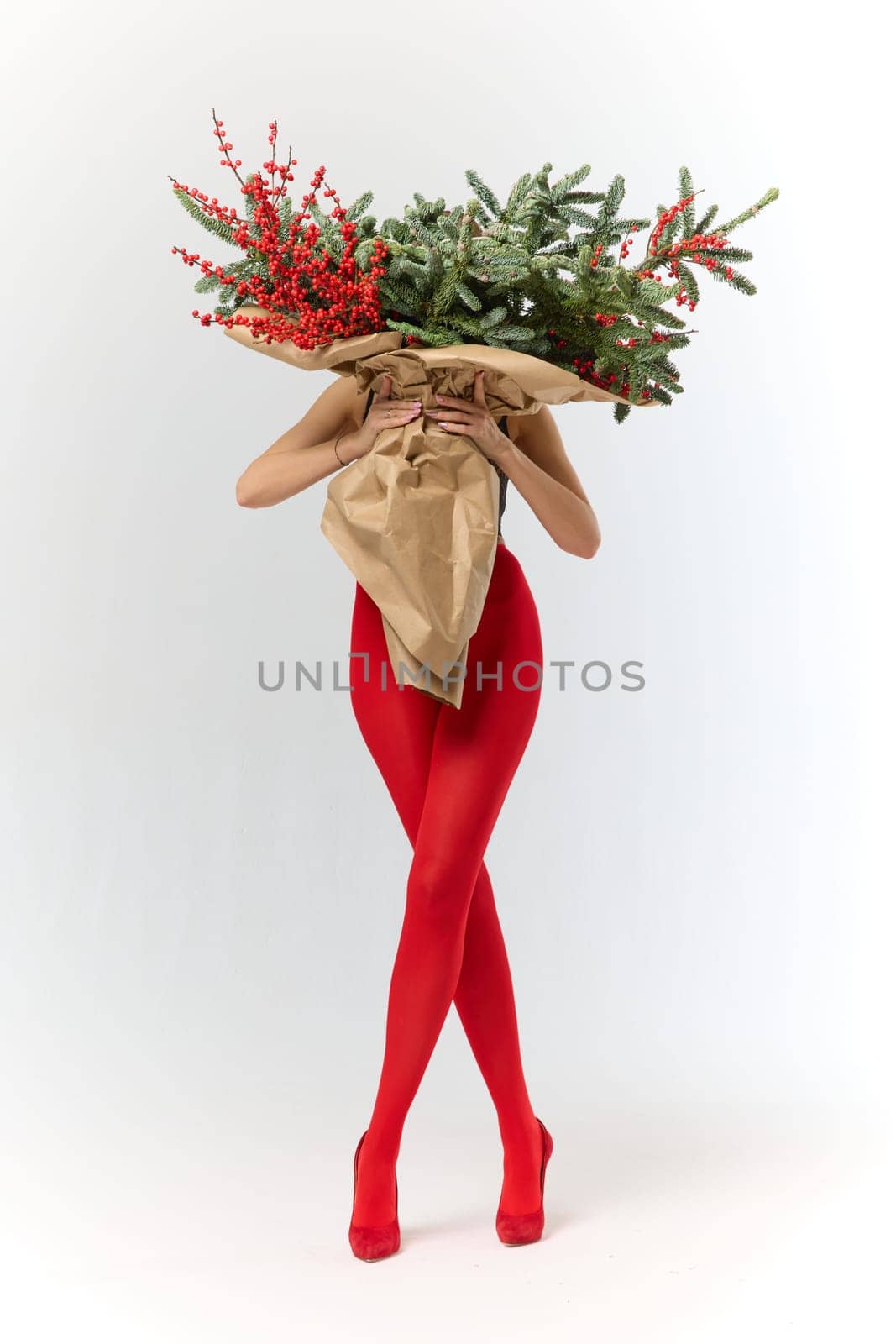 A long girl in red tights and high-heeled shoes holds a huge bouquet of fir branches and red berries wrapped in cardboard paper, she closes with a bouquet, photo studio with white background. High quality 4k footage