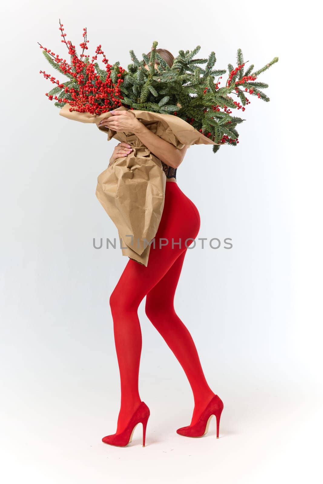 A long girl in red tights and high-heeled shoes holds a huge bouquet of fir branches and red berries wrapped in cardboard paper, she closes with a bouquet, photo studio with white background by vladimirdrozdin