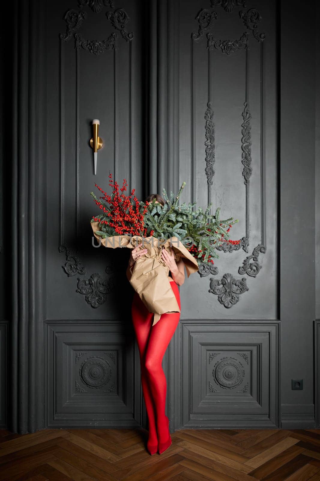 A long girl in red tights and high-heeled shoes holds a huge bouquet of fir branches and red berries wrapped in cardboard paper, she closes with a bouquet, photo studio with grey wall background. High quality 4k footage