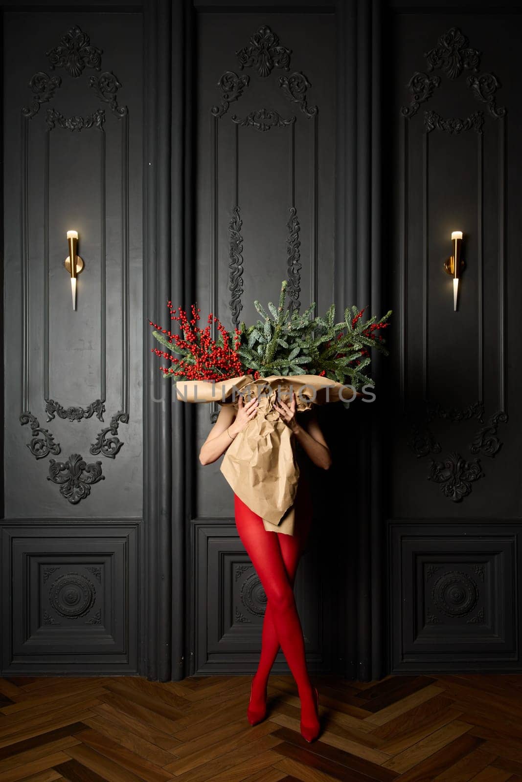 A long girl in red tights and high-heeled shoes holds a huge bouquet of fir branches and red berries wrapped in cardboard paper, she closes with a bouquet, photo studio with grey wall background by vladimirdrozdin