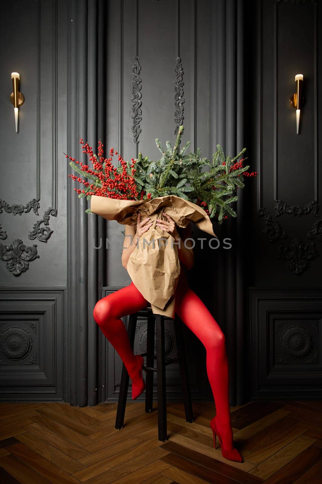 A long girl in red tights and high-heeled shoes holds a huge bouquet of fir branches and red berries wrapped in cardboard paper, she closes with a bouquet, photo studio with grey wall background by vladimirdrozdin