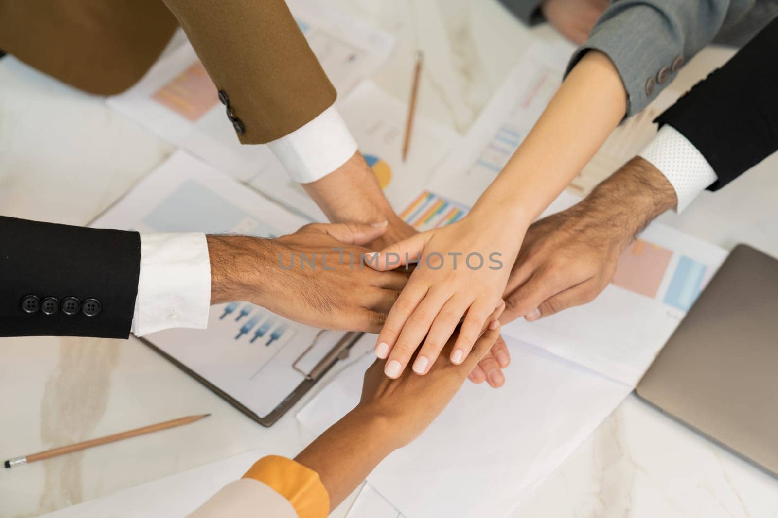 Top view of corporate diverse businesspeople putting hands together on meeting table with document scatter around at business meeting room. Represented unity, cooperation, collaboration. Ornamented.