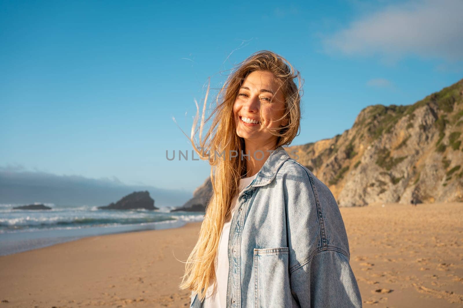 Portrait of smiling woman with long blond hair on sandy beach. Happy female in denim shirt with sea and mountain in background. Lady enjoying vacation against blue sky.