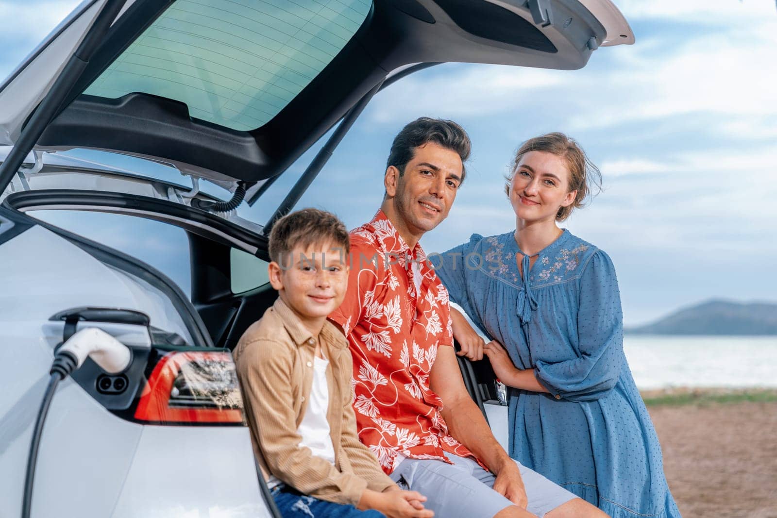 Family vacation trip traveling by the beach with electric car, lovely family sit on the trunk, charging EV car battery with green and sustainable energy. Family travel and eco-friendly car. Perpetual